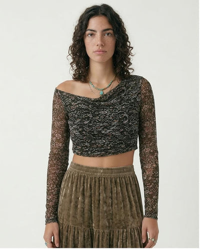 Woman wearing a black and gold patterned top with sheer sleeves and a brown pleated skirt on a white background