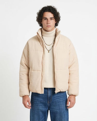 Man wearing a beige puffer jacket over a cream sweater with blue jeans on a white background