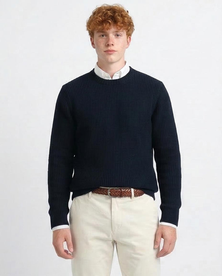 Man wearing a navy sweater over a white shirt with beige pants on a light gray background
