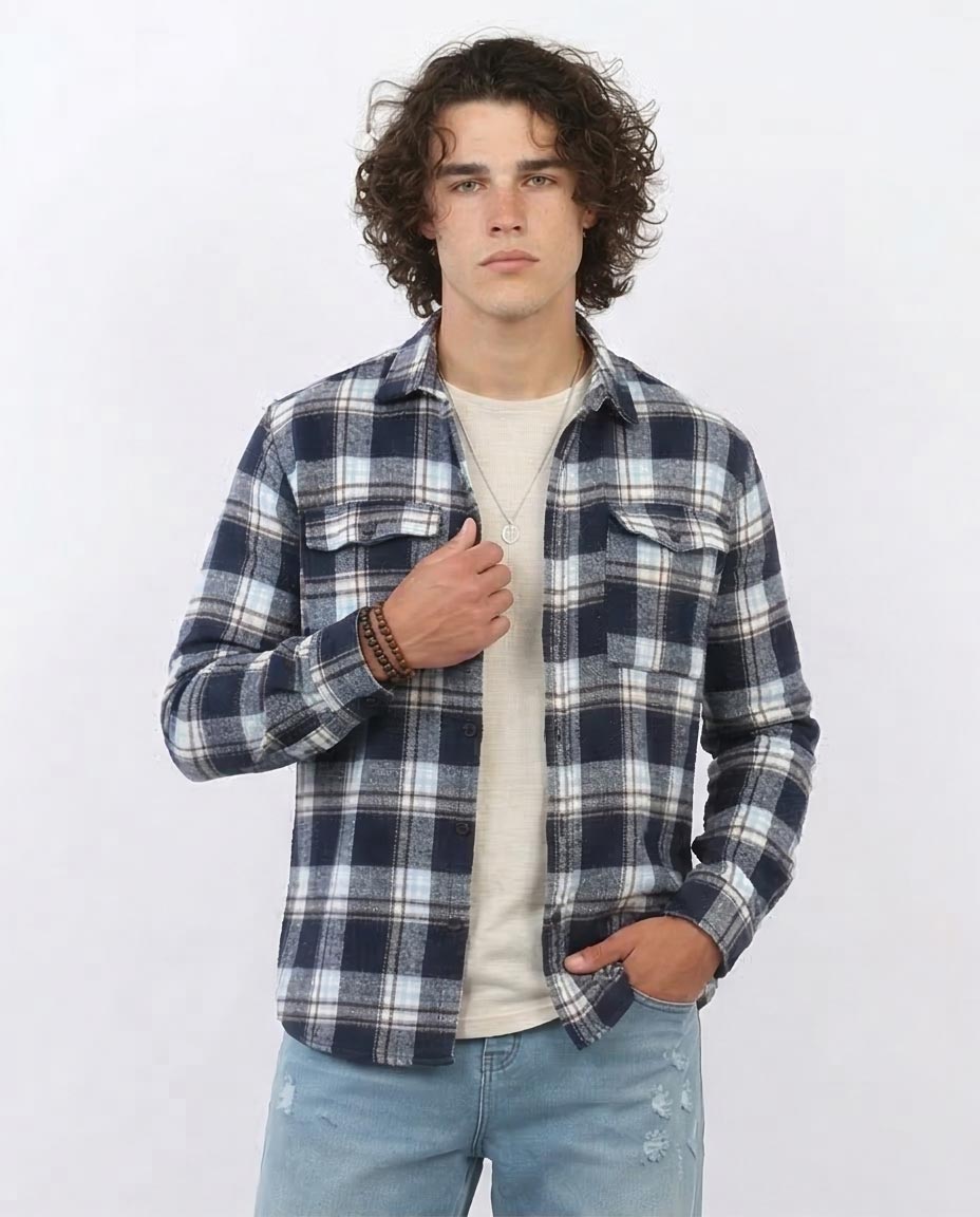 Man wearing a blue and white plaid shirt over a beige sweater with light blue jeans on a white background