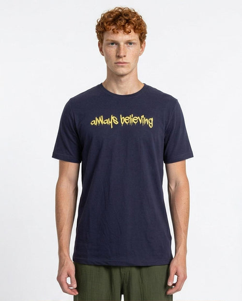 Man wearing a navy t-shirt with 'always believing' in yellow text on a white background