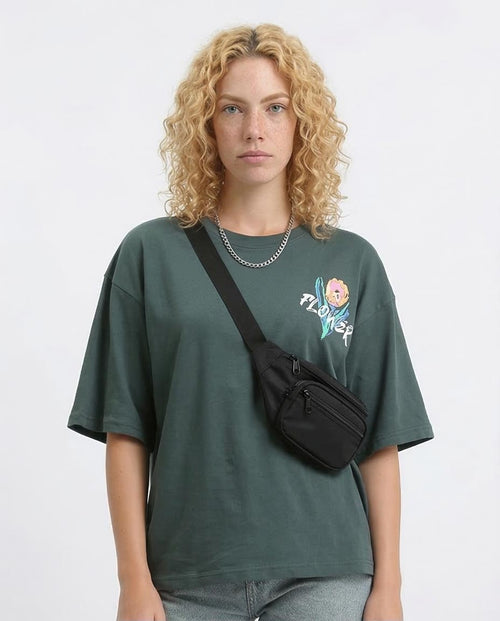 Person wearing a green t-shirt with a graphic design and carrying a black crossbody bag on a light gray background