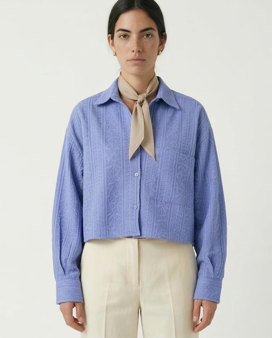 Woman wearing a blue textured shirt with a beige scarf and white pants on a light gray background