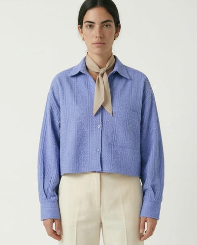 Woman wearing a blue textured shirt with a beige scarf and white pants on a light gray background