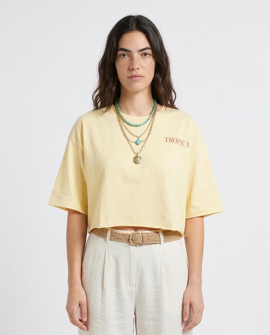 Woman wearing a yellow t-shirt with 'Tropical' text, white pants, and multiple necklaces on a light gray background