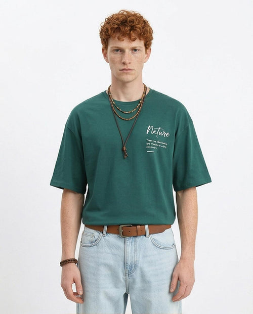 Man wearing a green t-shirt with text, light blue jeans, and multiple necklaces on a white background