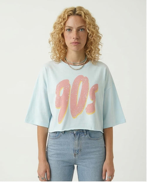 Person wearing a light blue t-shirt with '90s' text in pink and yellow on a plain background
