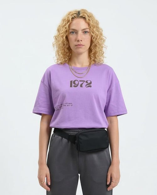 Woman wearing a purple t-shirt with '1972' text and gray pants on a white background