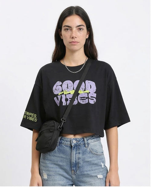 Person wearing a black t-shirt with 'Good Vibes' text, holding a black crossbody bag on a white background