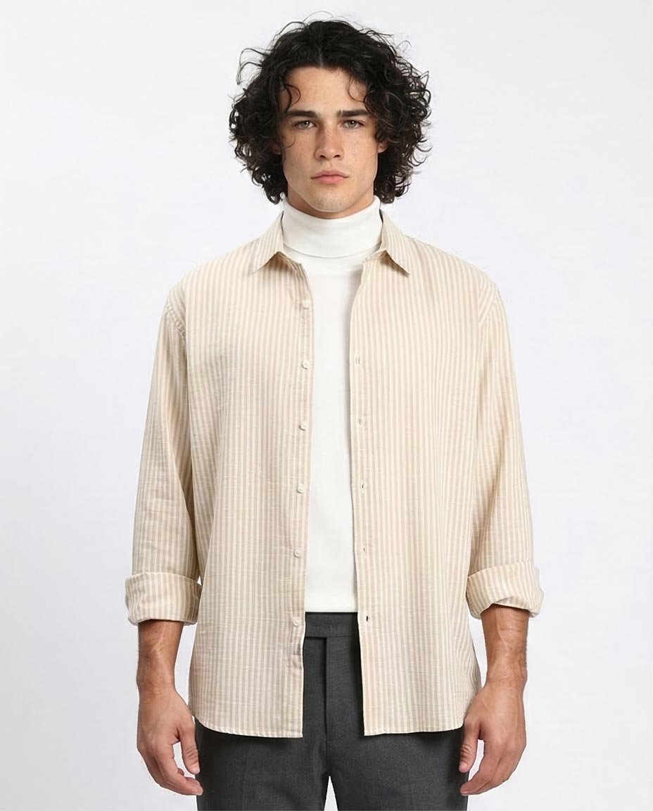 Man wearing a beige striped shirt over a white turtleneck with dark pants on a light gray background