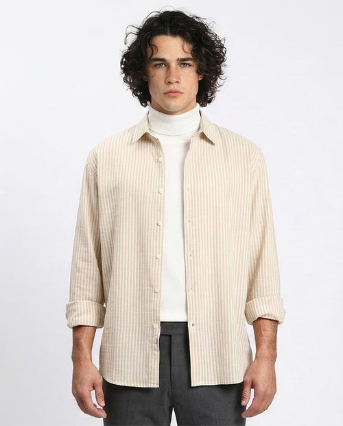 Man wearing a beige striped shirt over a white turtleneck with dark pants on a light gray background