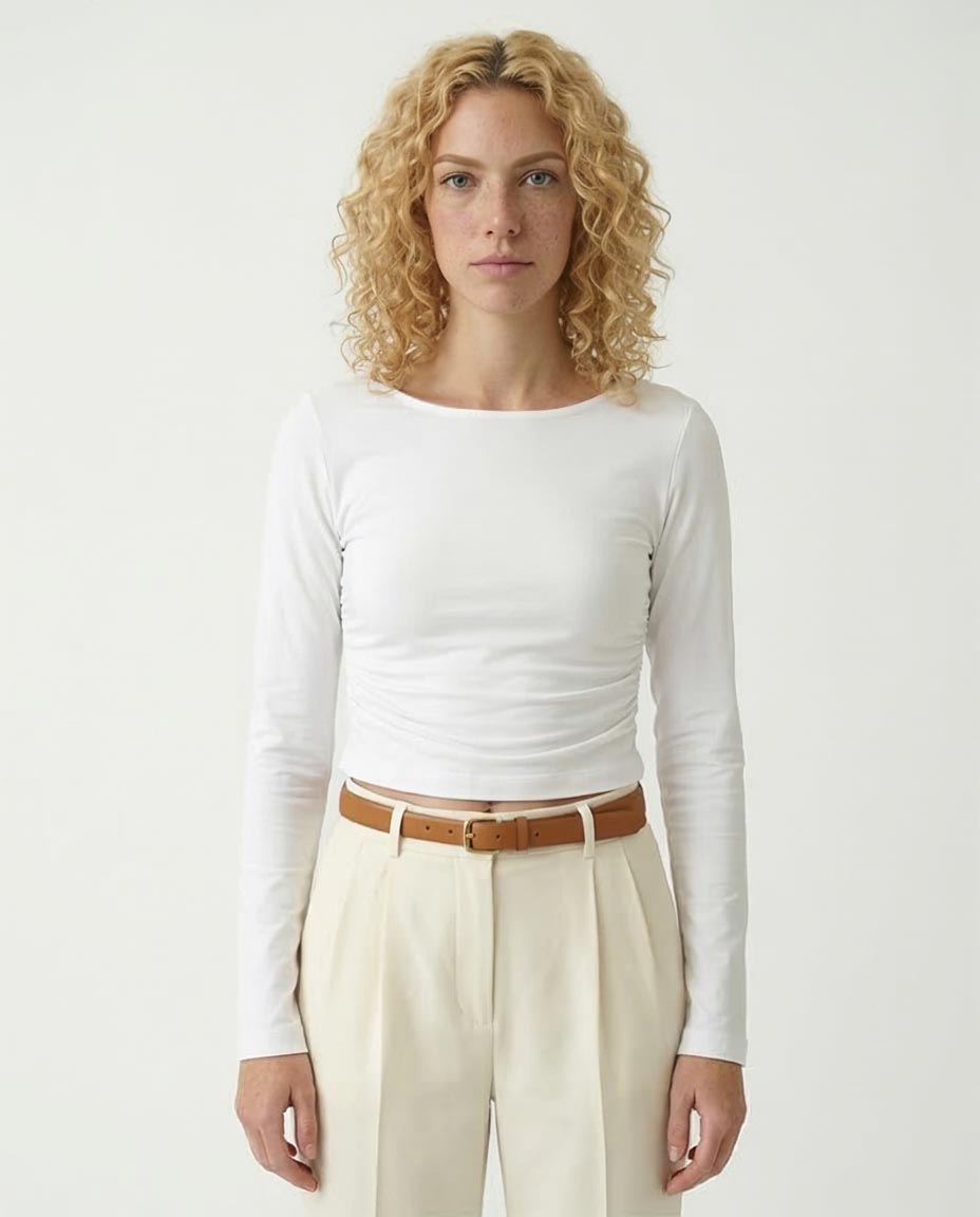 Woman wearing a white long-sleeve top and beige pants with a brown belt on a plain background