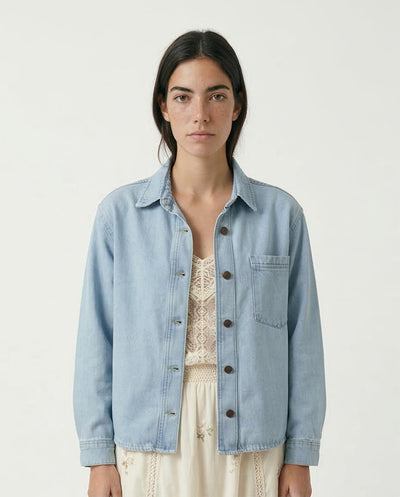 Woman wearing a light blue denim jacket over a white lace dress on a plain background